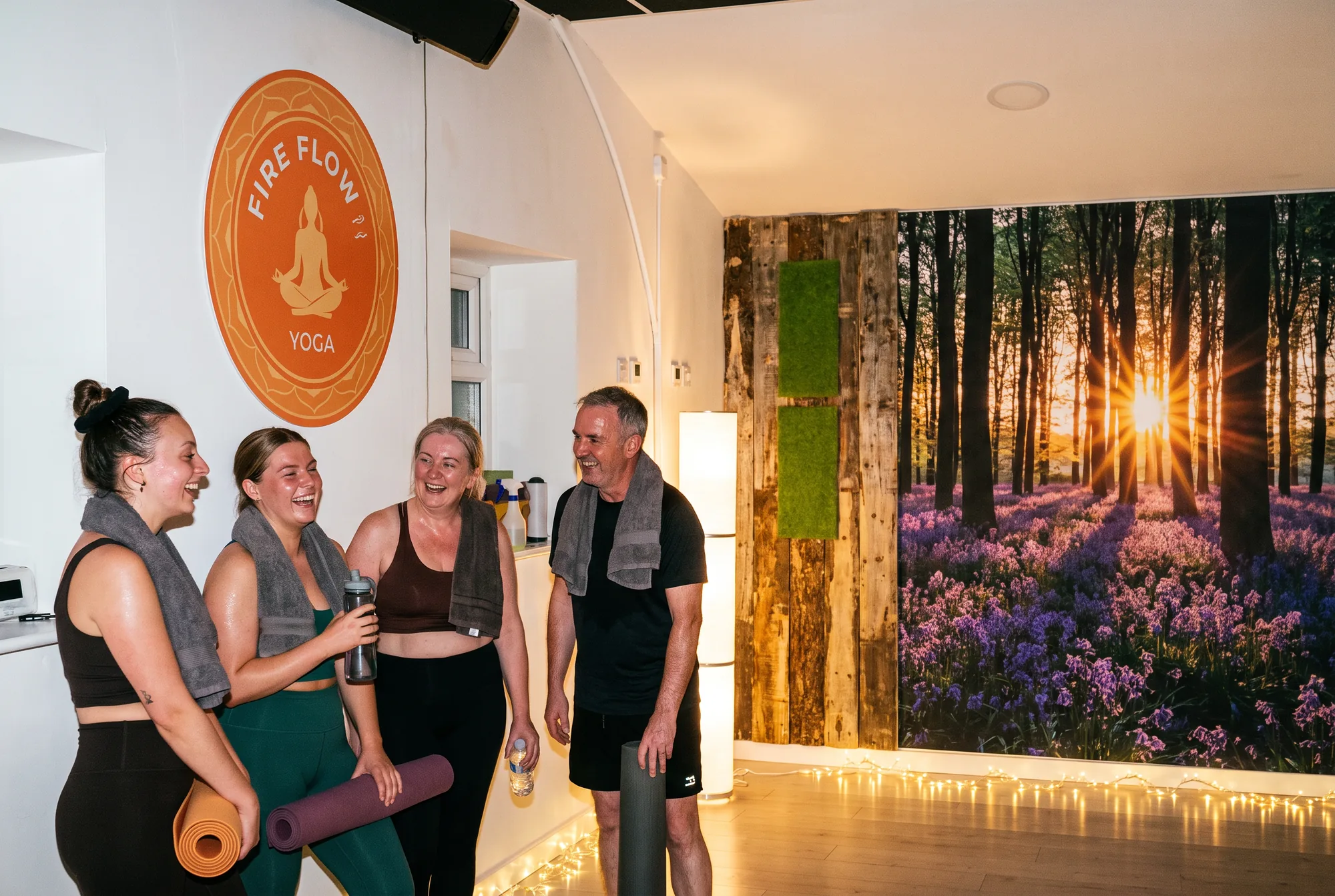 Four women and a man laughing after class with towels over their shoulders, the round orange Fire Flow Yoga sign on the wall behind them.