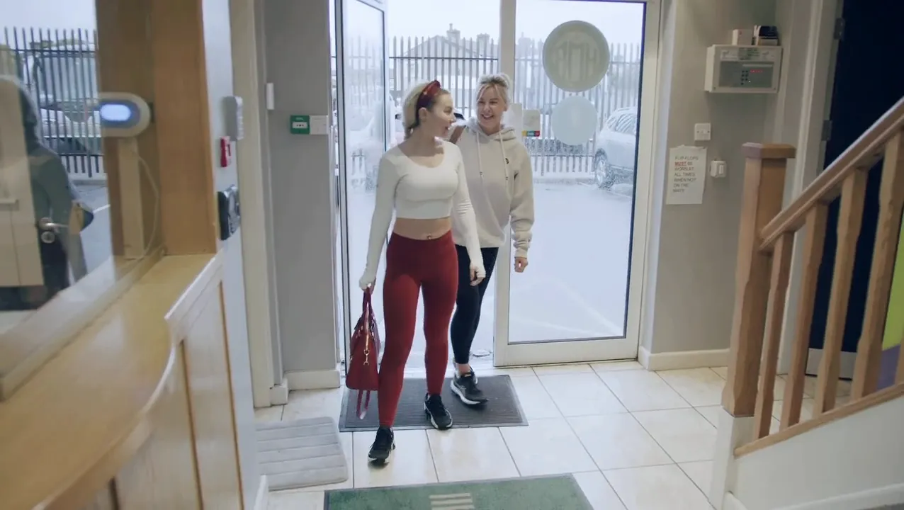 Two guests walk in through the front door of the building towards reception.